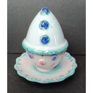 Puede incluir: Un vaso para huevos de cerámica con una cara caprichosa y un sombrero. El vaso está pintado en tonos de azul, verde y rosa.