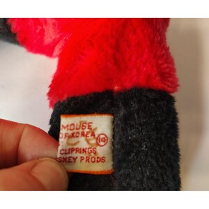 May include: A vintage plush toy featuring red and black fur. The attached tag reads "MOUSE OF KOREA" and "CLIPPINGS SNEY PRODS". The image focuses on the toy's fur and the tag. The toy is partially visible.