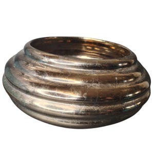 Op de afbeelding: Een metallic zilverkleurige kom met een brede, afgeronde vorm. De kom heeft een reeks horizontale, geribbelde banden rond de buitenkant. Het oppervlak heeft een licht verouderd uiterlijk, met subtiele kleur- en textuurvariaties. De kom is waarschijnlijk decoratief.