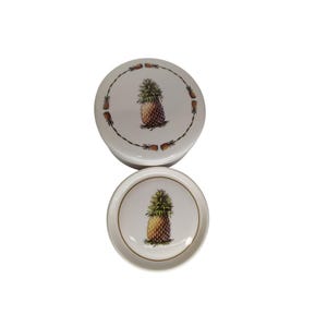 May include: Two white ceramic round containers with lids. Both containers feature a pineapple design with brown and green details. The larger container is on top of the smaller container.