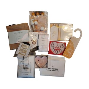 Puede incluir: Una colección de artículos temáticos de boda, que incluyen una bolsa de yute, artículos de tocador de viaje, etiquetas de regalo, un soporte para anillos en forma de corazón y un colgador de puerta. Los artículos están dispuestos sobre una superficie blanca, con el texto "Bride to Be".