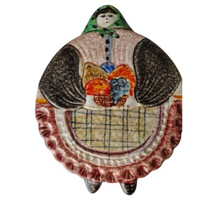 May include: A ceramic figurine in the shape of a woman, wearing a green headscarf, pink blouse, and black sleeves. The figure holds a basket of colorful fruit and mushrooms. The apron has a plaid pattern, and the bottom edge is scalloped.