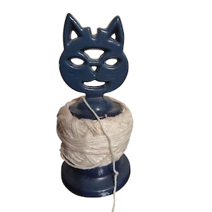 Puede incluir: Un soporte para hilo en forma de gato azul oscuro con una madeja de hilo blanco. La cara del gato tiene ojos recortados y una boca sonriente. El hilo está enrollado alrededor de la base de la figura del gato.