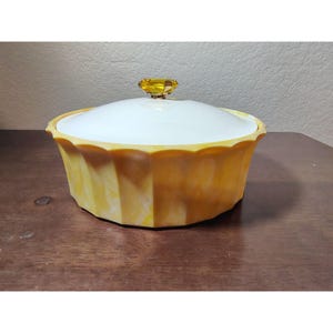 Könnte beinhalten: Eine Vintage-Servierschüssel mit Deckel in Gelb und Weiß. Die Schüssel hat einen gewellten Rand und ein facettiertes Design. Der Deckel ist weiß mit einem gelben, edelsteinartigen Griff. Die Schüssel steht auf einer Holzoberfläche.