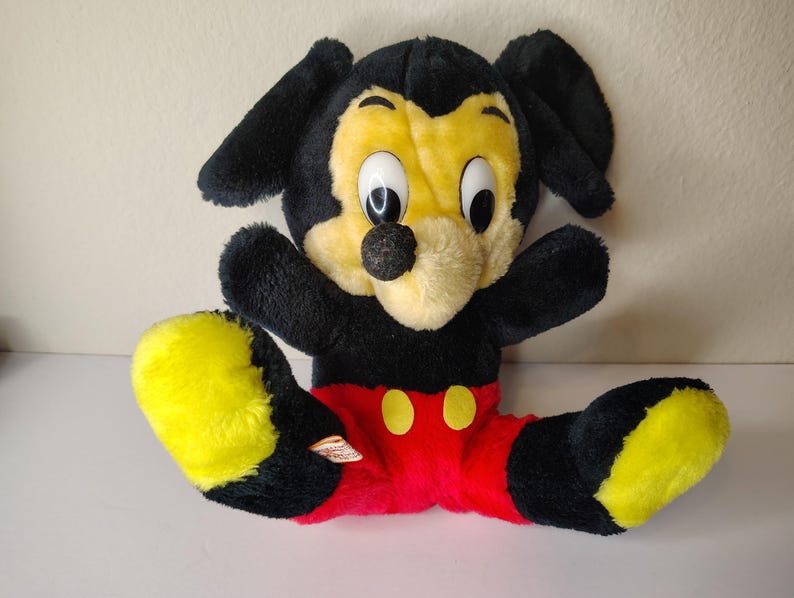 May include: A vintage Mickey Mouse soft toy. The toy has a black head and ears, a yellow face, and a black nose. It is wearing red shorts with yellow buttons and has yellow feet. The toy is in a sitting position with its arms outstretched.