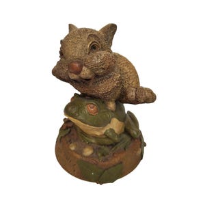 May include: A brown rabbit figurine perched on top of a green frog figurine. Both figurines are sitting on a brown base with green leaves.
