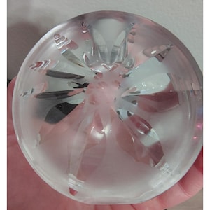 May include: A clear, round glass paperweight with a frosted finish. The paperweight features a starburst design with pointed, cut facets radiating from the center. The facets catch and reflect light, creating a sparkling effect. The paperweight is held in a hand.