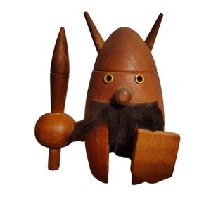 Puede incluir: Figura de vikingo de madera con acabado marrón. La figura tiene un casco con cuernos, una barba negra y un escudo. Sostiene una lanza. Los ojos están acentuados con anillos dorados. Un artículo decorativo.