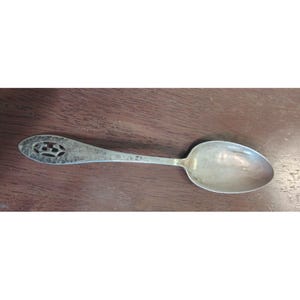 Puede incluir: Una cuchara de metal plateado con un acabado martillado y texturizado en el mango. El extremo del mango incluye un diseño calado con un emblema central. La parte cóncava de la cuchara es lisa, de forma ovalada y refleja la luz.