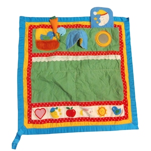 May include: A square fabric activity mat featuring a green polka dot center, bordered by red and yellow fabric. It includes attached plush elements like a carrot, a blue bird, a moon, a mirror, and a bottom row of icons including a heart, apple, and sun.
