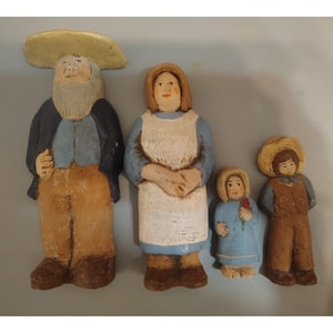 May include: A set of four hand-painted figurines depicting a family. The group includes a tall figure with a wide-brimmed hat, a figure in a blue dress and white apron, a small child in a blue dress holding a red flower, and a child in a straw hat.