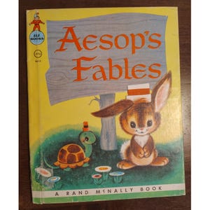 May include: A vintage children's book titled Aesop's Fables with a yellow cover. The illustration features a brown rabbit wearing a small hat and a green turtle with a brown shell. A gray wooden sign is in the background with the book title.