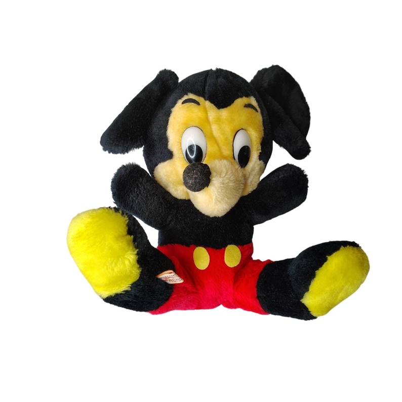 May include: Vintage Mickey Mouse soft toy. The plush has a black body, ears, and arms, with a yellow face and feet. Mickey is wearing red shorts with yellow buttons. The toy is in a sitting position with its legs spread.