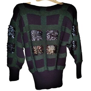 May include: A vintage sweater with a green and dark purple plaid pattern. The sweater features square sequin embellishments and a black ribbed hem and cuffs. The sweater is hanging on a wooden hanger.