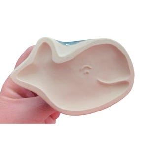 May include: A ceramic whale-shaped dish with a light pink interior and a blue top. The dish has a simple, minimalist design, with a curved shape and a small, subtle detail on the side. The whale's mouth is slightly open.