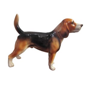 Könnte beinhalten: Eine Beagle-Figur aus Keramik, die im Profil mit erhobenem Schwanz dargestellt ist. Der Hund hat ein dreifarbiges Fell mit schwarzem Rücken, lohfarbenen Seiten und weißen Abzeichen an Schnauze, Brust und Pfoten. Die Oberfläche ist glatt und glänzend.