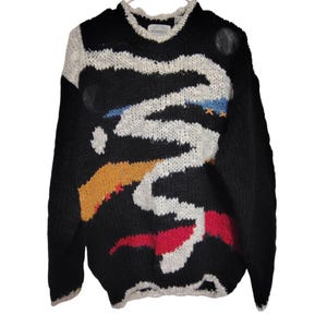 May include: A black long-sleeve knit sweater featuring a large, thick white abstract zigzag pattern across the front. The design includes horizontal accents in yellow, red, and blue, along with small circular black patches and orange cross-stitch details.
