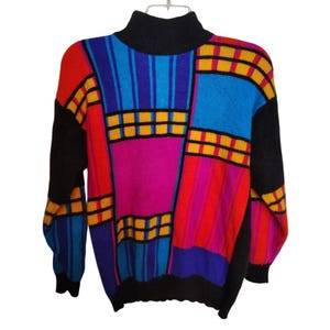 Może przedstawiać: Kolorowy sweter z czarnym golfem i rękawami. Sweter ma wzór patchwork z czerwonymi, niebieskimi, żółtymi i fioletowymi geometrycznymi kształtami. Sweter jest wykonany z miękkiego materiału.