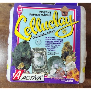 Puede incluir: Un paquete de Celluclay papel maché instantáneo gris original. El paquete tiene un diseño morado y azul con el nombre de la marca en amarillo. Incluye una hoja de proyecto y pesa 454 g.