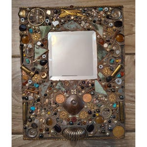 Puede incluir: Un espejo rectangular con un marco de mosaico. El marco está adornado con una variedad de objetos pequeños, incluyendo cuentas, monedas y adornos metálicos en tonos marrones, dorados y turquesas. El espejo tiene un área de visualización cuadrada.