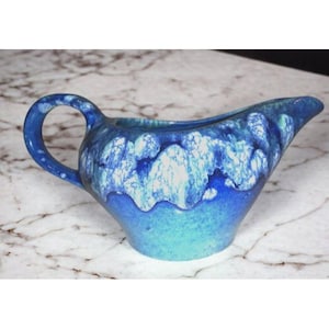 Könnte beinhalten: Eine blaue Keramik-Sauciere mit Henkel. Die Sauciere hat ein einzigartiges Design mit einer Mischung aus blauen und weißen Farben. Der Griff hat eine Schlaufenform und der Ausguss ist spitz. Die Sauciere ist wahrscheinlich Vintage.