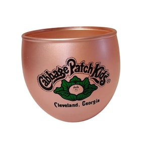 May include: A rose gold-coloured, rounded glass featuring the Cabbage Patch Kids logo in black and green. The logo includes a cabbage with a smiling face and the text "Cleveland, Georgia."