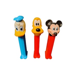 Peut inclure: Trois distributeurs PEZ vintage représentant des personnages Disney : Donald Duck, Pluto et Mickey Mouse. Donald Duck a un chapeau et un bec bleus, Pluto est jaune et Mickey Mouse a des oreilles noires. Chaque distributeur a un corps en plastique coloré.