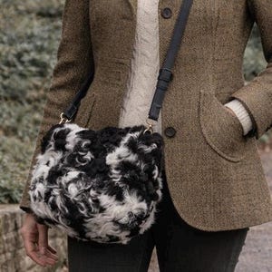 May include: A black and white fluffy crossbody bag with a black leather strap. The bag has a soft, textured appearance and is worn over the shoulder. The bag is paired with a brown tweed jacket.