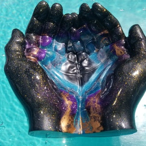 May include: A pair of resin hands cupped together, forming a bowl. The hands are painted black with gold and purple glitter, and the inside of the bowl is filled with a blue, purple, and gold resin mixture.