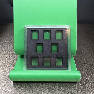 May include: A bright green plastic display stand with a black insert featuring nine square openings. The stand has a curved base and a vertical back, designed to hold and showcase small items or collectibles.