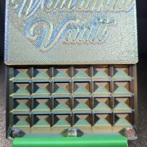 May include: A 3D-printed box with the text "Valcambi Vault" in a script font. The box has a textured surface with a geometric pattern and a green cylindrical handle. The colors include gold, green, and blue.