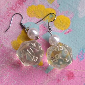 May include: A pair of dangle earrings featuring a clear, iridescent, multi-sided dice charm with white numbers and a white pearl bead above each dice. The earrings have silver hooks.