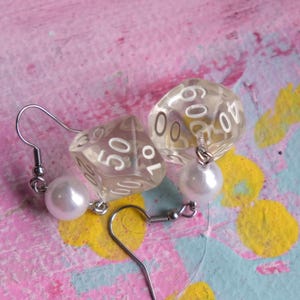 May include: A pair of earrings featuring clear plastic dice with white numbers and a white pearl charm on each earring.