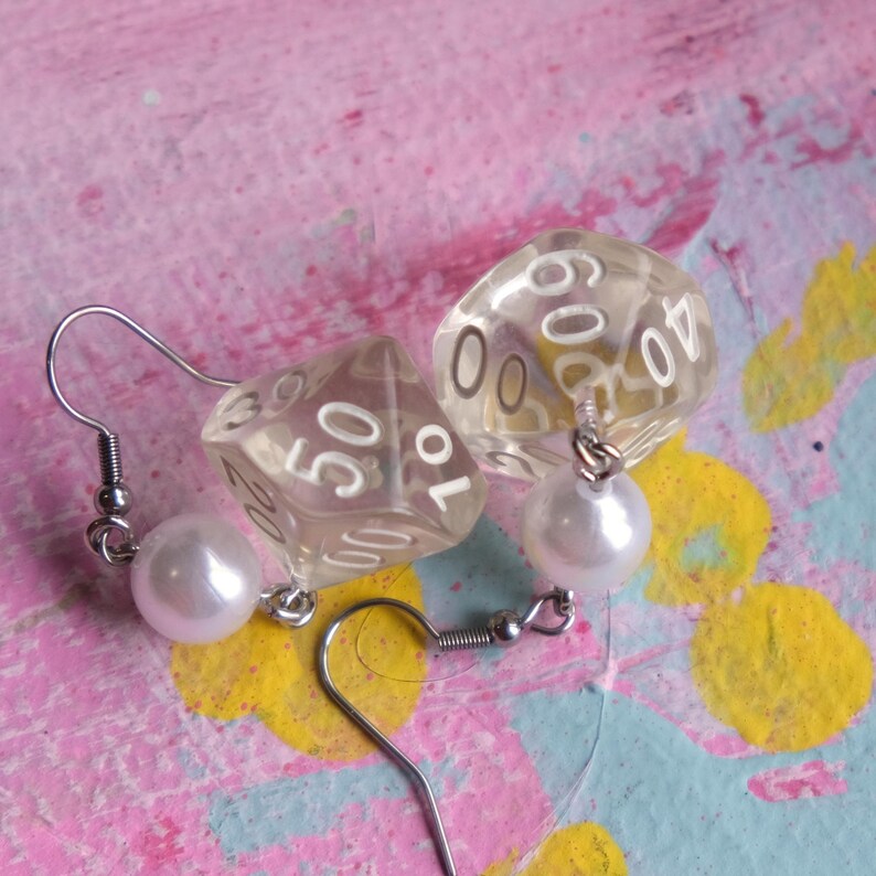 May include: A pair of earrings featuring clear plastic dice with white numbers and a white pearl charm on each earring.