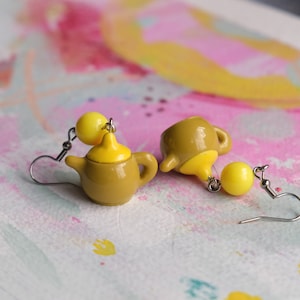 May include: A pair of earrings featuring miniature teapot charms with yellow lids and brown bodies. The earrings are attached to silver hooks with yellow bead accents.
