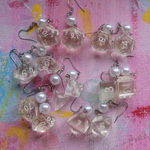 May include: A set of 10 clear plastic earrings featuring a pearl charm and a dice-shaped charm with white numbers. The dice charms are in various shapes and sizes.