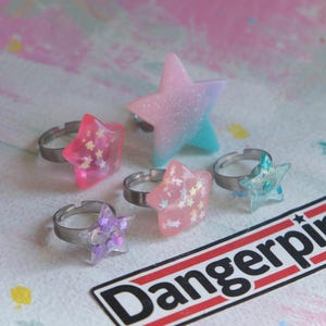 May include: A selection of star-shaped rings in shades of pink, purple, and blue, with adjustable silver bands. Some stars feature glitter or star-shaped details. The rings are presented on a vibrant, patterned surface.