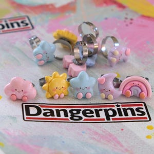 May include: A collection of colorful rings with cute, whimsical designs. The rings feature shapes like stars, clouds, suns, moons, and rainbows. Each ring has a silver band and is decorated with pastel colors and smiling faces. The word "Dangerpins" is printed on a white sticker.