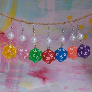 Puede incluir: Seis pares de pendientes con una perla y un dado de plástico transparente de 20 caras en diferentes colores: amarillo, rosa, verde, rojo, azul y morado.