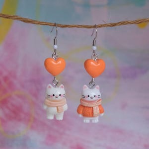 May include: Dangle earrings with a white cat charm wearing a scarf and an orange sweater, hanging below an orange heart. The earrings have silver-coloured hooks and a small white bead. The earrings are suspended from a string.
