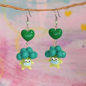 Può includere: Un paio di orecchini fatti a mano con un cuore verde e un personaggio stravagante, simile a un cartone animato, con la testa a forma di broccoli. Gli orecchini sono sospesi a un gancio color argento e una piccola perla verde.