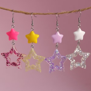 May include: Four pairs of star-shaped dangle earrings with glitter accents. The earrings are made of clear resin with a star-shaped cutout in the centre. Each pair features a different colour star charm: pink, yellow, purple, and white.