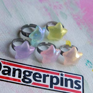 May include: Six star-shaped rings in various iridescent colors: blue, yellow, pink, and purple. The rings have adjustable silver bands. The word "Dangerpins" is visible on a sticker in the lower part of the image.
