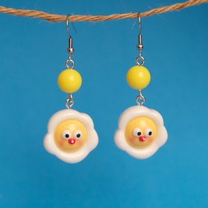 May include: A pair of earrings featuring a white and yellow fried egg design with a red nose and black eyes. The earrings are suspended from silver hooks with a yellow bead above each egg.