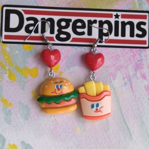 Op de afbeelding: Een paar hangende oorbellen met rode hartcharms. Eén oorbel heeft een gele en groene hamburger met een glimlachend gezicht. De andere oorbel heeft een gele en oranje frietbak met een glimlachend gezicht. De oorbellen zijn gelabeld met "Dangerpins".