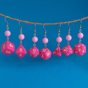 May include: Pink dice earrings with white numerals, suspended from a brown string against a blue backdrop. Each earring has a pink bead and a silver hook. The dice are various polyhedral shapes, perfect for a gamer.