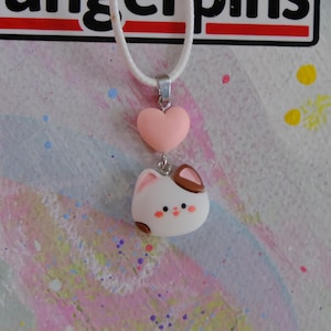 May include: A white cord necklace with a silver-tone pendant featuring a pink heart and a white cat-shaped charm with brown accents. The cat charm has rosy cheeks and small black dots for eyes and a nose.