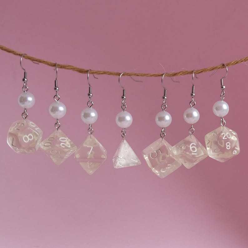 May include: Seven pairs of dangle earrings featuring clear dice charms and white pearl beads. The dice charms are numbered 1-6, 7, 8, 9, 10, 12, and 20.