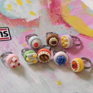 May include: A collection of nine adjustable metal rings featuring miniature food-themed charms made of polymer clay. The charms include various colorful cakes, tarts, and pastries with detailed textures, such as berries, cream, and chocolate toppings.