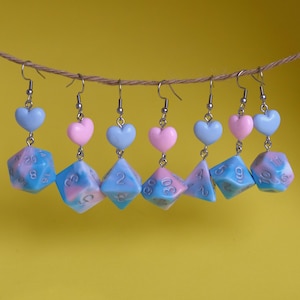 May include: Six pairs of dangle earrings featuring a blue and pink swirl dice charm with a heart charm above it. The dice charms have silver numbers on them.